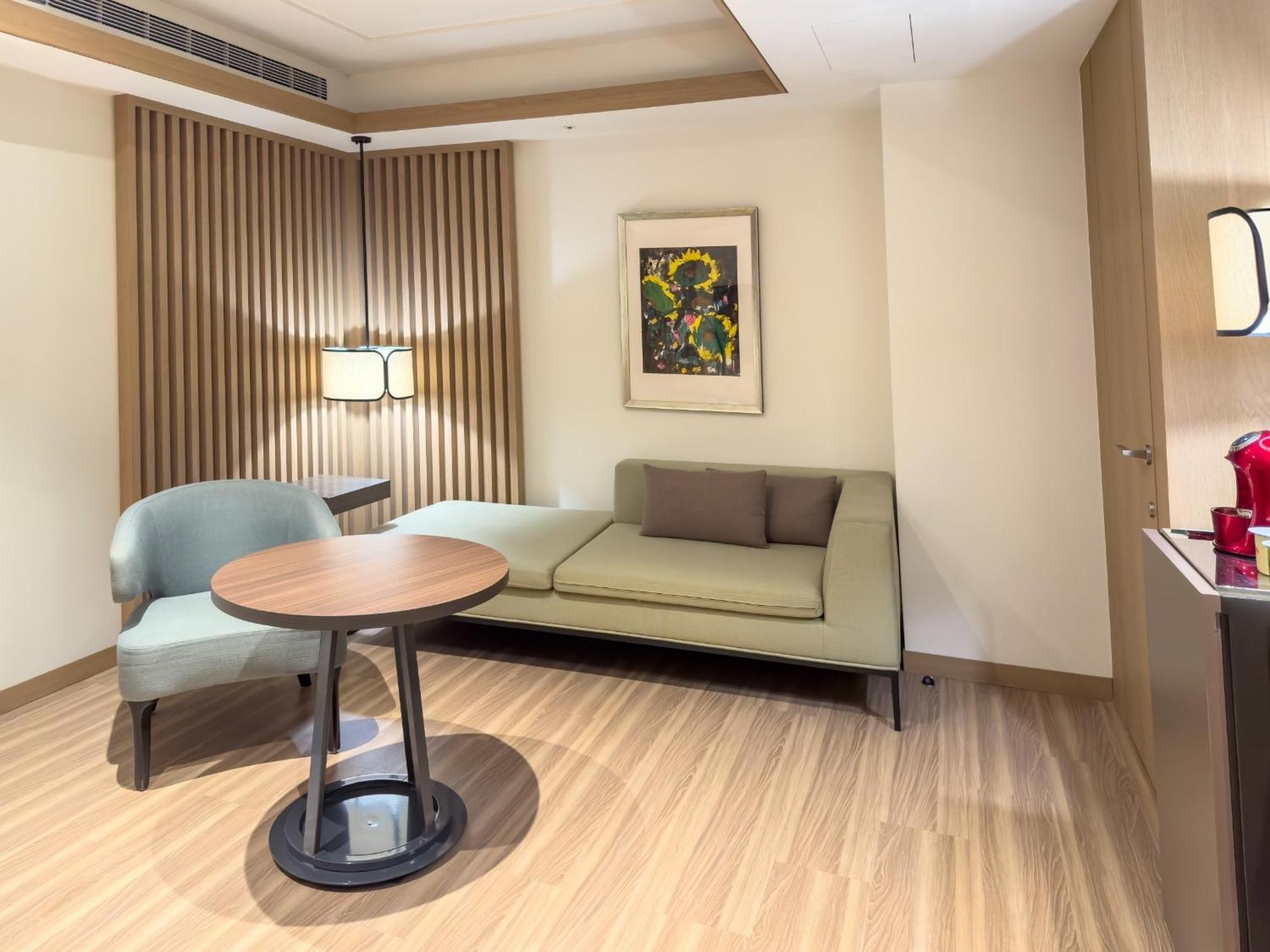 فندق سيزار مترو تايبيه المظهر الخارجي الصورة The photo shows a cozy interior space, likely a hotel room or a living area. On the left side, there’s a light green sofa with two pillows and a small round table in front of it. To the left of the sofa, there's a stylish chair. A piece of artwork is