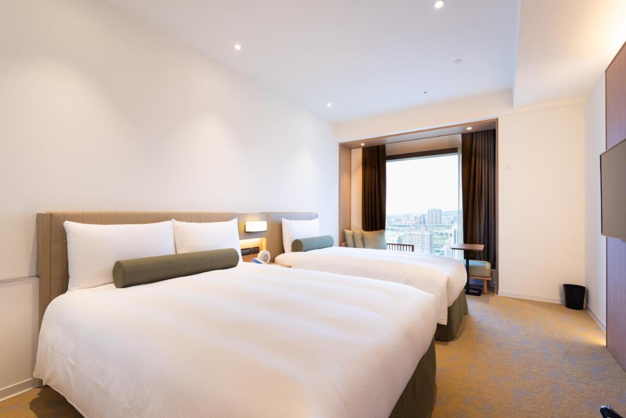 فندق سيزار مترو تايبيه المظهر الخارجي الصورة The photo shows a modern hotel room with two beds. Each bed has crisp white linens and a decorative gray bolster pillow. The room features a minimalist design with light-colored walls and a neutral carpet. There’s a large window with curtains that pa
