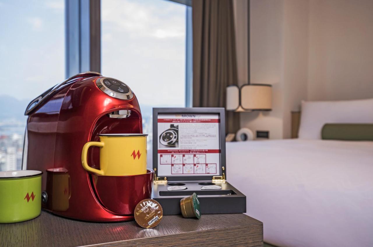 فندق سيزار مترو تايبيه المظهر الخارجي الصورة The image shows a cozy hotel room setting featuring a red coffee maker on a wooden table. Next to the coffee maker are two yellow mugs and a brown capsule, likely used for coffee. There is also a box containing a selection of coffee capsules. In the 