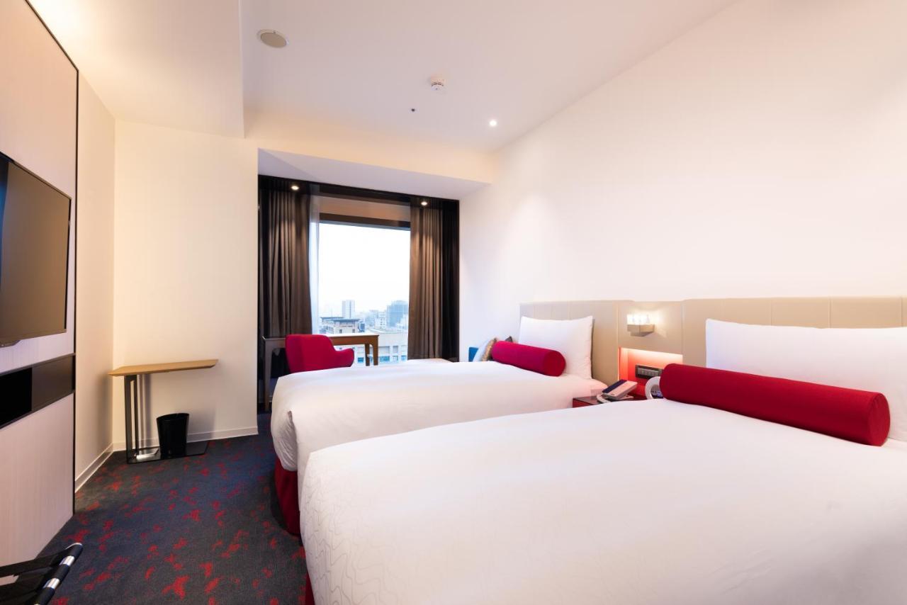فندق سيزار مترو تايبيه المظهر الخارجي الصورة The photo shows a modern hotel room featuring two beds with white bedding and red accent pillows. There is a small wooden table next to a window that has dark curtains, allowing for natural light to enter the room. The walls are painted in light colo