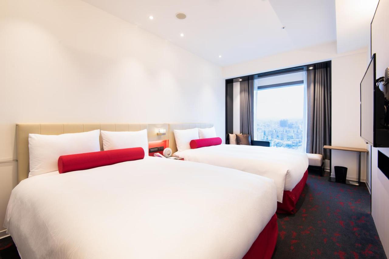 فندق سيزار مترو تايبيه المظهر الخارجي الصورة The image shows a modern hotel room featuring two queen-sized beds with white bedding and red accents. The room is spacious with neutral-colored walls, and there is a large window that provides natural light and a view. On one side, there's a small d