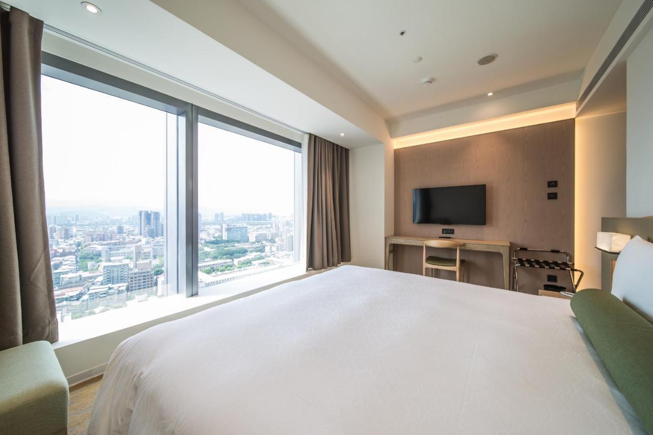 فندق سيزار مترو تايبيه المظهر الخارجي الصورة The photo shows a modern hotel room featuring a large bed with a white bedspread. There is a large window that offers a view of the city skyline, allowing natural light to fill the space. To the side, there is a flat-screen TV mounted on the wall, an