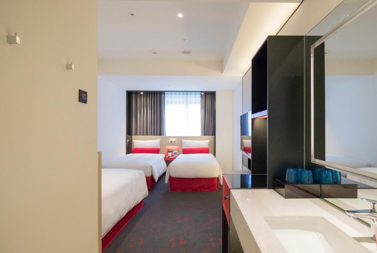 فندق سيزار مترو تايبيه المظهر الخارجي الصورة The photo shows the interior of a hotel room. It features two single beds with white linens and red accents. There is a window with sheer curtains allowing natural light to enter the room. The floor is carpeted, and the decor is modern and minimalist